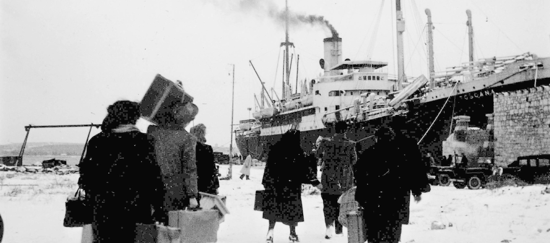 Un gruppo di persone di spalle cammina verso una grande nave ormeggiata. Le persone portano con sé valigie e bagagli, e la scena è ambientata in un ambiente invernale, con neve a terra. La nave, visibile sulla destra, emette fumo dal camino e appare pronta a partire.