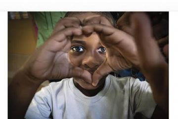 L'immagine è dominata da una fotografia a colori in primo piano che ritrae il volto di un bambino, il quale guarda dritto verso l'obiettivo e accosta le mani davanti agli occhi, unendo i pollici e gli indici per formare la sagoma di un cuore. Così risaltano i suoi occhi, espressivi e luminosi. Indossa una maglietta bianca. Subito sotto la foto, spicca il titolo della mostra "SONRISAS Y ESPERANZA" scritto in lettere maiuscole e di un colore giallo acceso. Quindi, sotto il titolo principale, in nero e con un carattere più piccolo, si legge: "mostra fotografica di Paolo Rocchi". Poco sotto, su sfondo bianco, sono riportati i dettagli dei due eventi speciali: l'inaugurazione con aperitivo del 6 giugno e il reading poetico di Simona Giorgi del 13 giugno. A destra, separato da una sottile linea verticale nera, c'è il blocchetto con tutti gli orari di apertura dei fine settimana e la specifica "INGRESSO GRATUITO" in grassetto. Ancora sotto, ma al centro, scritto in grande (le date in giallo e il luogo in nero), si trovano le informazioni principali: "DAL 6 AL 21 GIUGNO 2026", "ORATORIO DEI BIANCHI" e "CENTRO STORICO CASTELNUOVO MAGRA (SP)". In fondo alla pagina, una striscia orizzontale mostra i loghi dei patrocini istituzionali (tra cui il Comune di Castelnuovo Magra e la Bandiera Arancione del Touring Club) e dei partner/sponsor dell'evento.