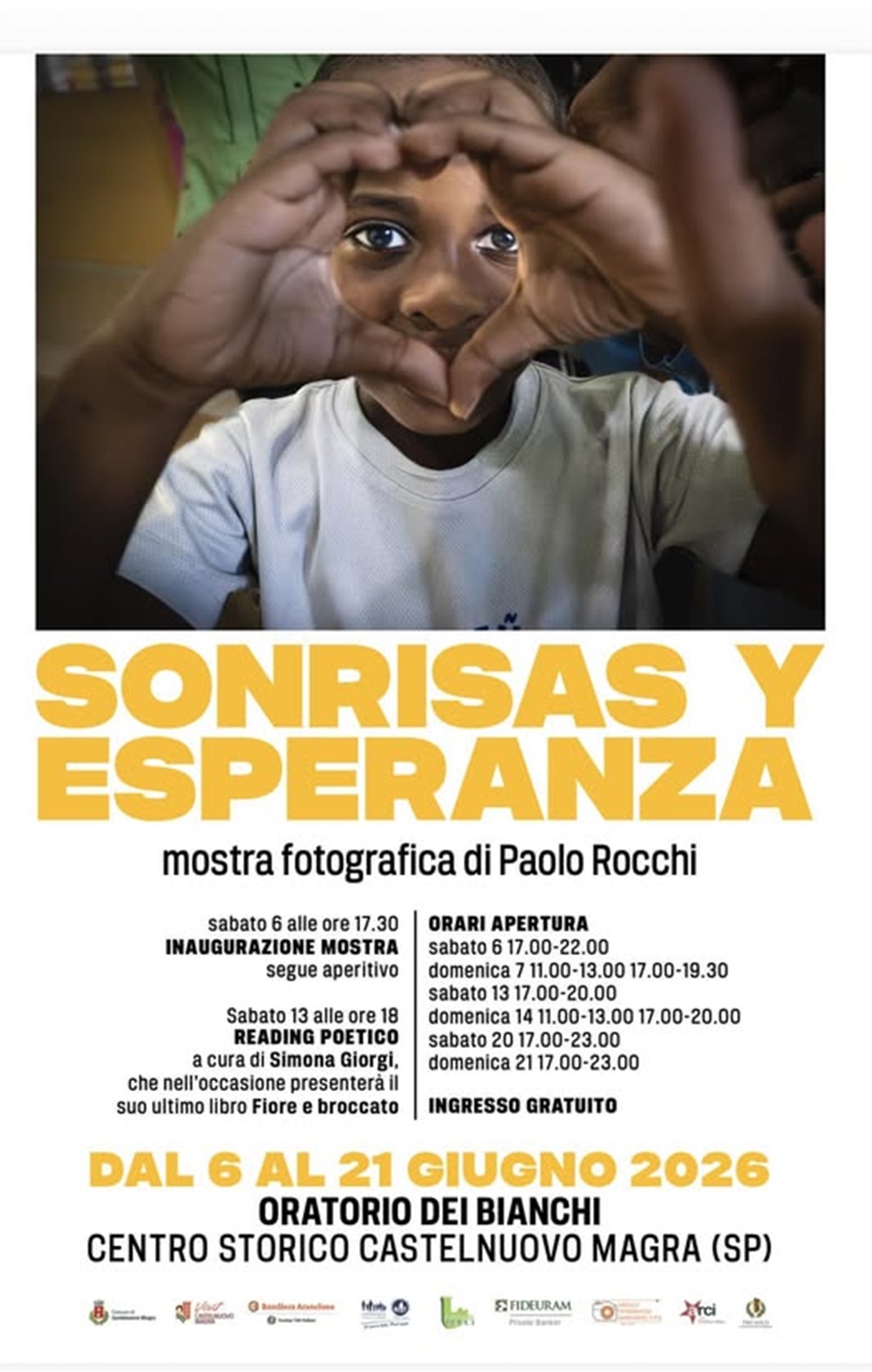 L'immagine è dominata da una fotografia a colori in primo piano che ritrae il volto di un bambino, il quale guarda dritto verso l'obiettivo e accosta le mani davanti agli occhi, unendo i pollici e gli indici per formare la sagoma di un cuore. Così risaltano i suoi occhi, espressivi e luminosi. Indossa una maglietta bianca. Subito sotto la foto, spicca il titolo della mostra "SONRISAS Y ESPERANZA" scritto in lettere maiuscole e di un colore giallo acceso. Quindi, sotto il titolo principale, in nero e con un carattere più piccolo, si legge: "mostra fotografica di Paolo Rocchi". Poco sotto, su sfondo bianco, sono riportati i dettagli dei due eventi speciali: l'inaugurazione con aperitivo del 6 giugno e il reading poetico di Simona Giorgi del 13 giugno. A destra, separato da una sottile linea verticale nera, c'è il blocchetto con tutti gli orari di apertura dei fine settimana e la specifica "INGRESSO GRATUITO" in grassetto. Ancora sotto, ma al centro, scritto in grande (le date in giallo e il luogo in nero), si trovano le informazioni principali: "DAL 6 AL 21 GIUGNO 2026", "ORATORIO DEI BIANCHI" e "CENTRO STORICO CASTELNUOVO MAGRA (SP)". In fondo alla pagina, una striscia orizzontale mostra i loghi dei patrocini istituzionali (tra cui il Comune di Castelnuovo Magra e la Bandiera Arancione del Touring Club) e dei partner/sponsor dell'evento.